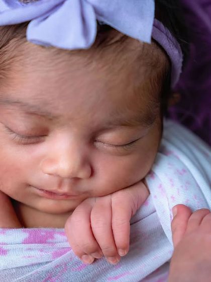 A close-up of a newborn girl with a purple bow, sleeping peacefully on a matching purple blanket.
