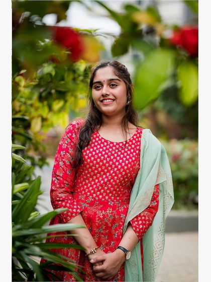 A lovely outdoor portrait of a young woman in a red and green salwar kameez, surrounded by lush greenery.