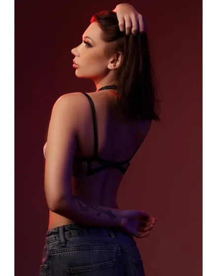 A moody portrait of a model in a harness bra and jeans, lit with a dramatic red light from the side that creates deep shadows and a sense of intimacy.