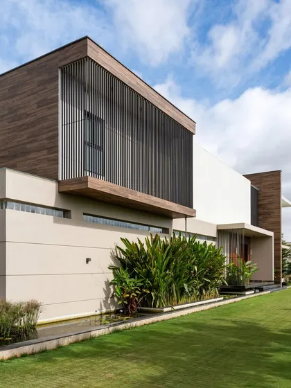 The home's exterior features strong geometric forms, with slatted screens providing both privacy and visual texture. The structure is designed to sit harmoniously within the surrounding mango plantations and cultivated land.