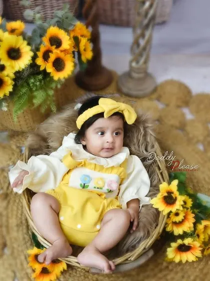 A lovely portrait of a baby girl in a yellow outfit and matching bow headband, sitting in a rustic basket. The sunflower props add a bright and cheerful element to the photo.