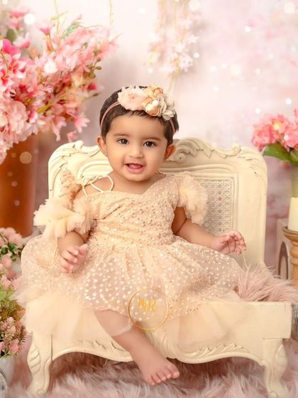A baby girl sits on a miniature throne in a lavish pink floral setting, looking like a little princess.