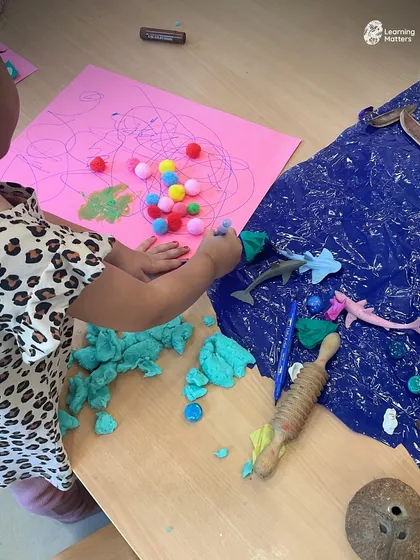 An invitation to create with multiple textures at once. Playdough, pom-poms, and an ocean-themed sensory mat allow this child to build a multi-layered story, combining different materials to express their ideas.