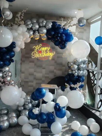 An elegant birthday setup with a circular balloon arch in navy blue, chrome silver, and white. The 'Happy Birthday' neon sign adds a warm glow to the celebration.