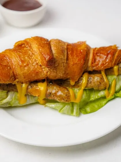 A croissant filled with sausage, lettuce, and a drizzle of mustard, shot on a clean white plate. This image is perfect for a cafe menu, showcasing a savory breakfast or lunch item.