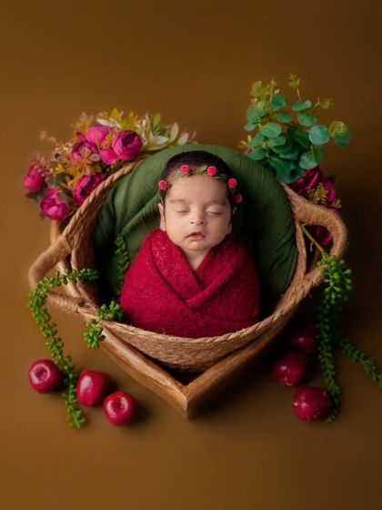 A newborn wrapped in red sleeps in a basket decorated with green leaves and bright red cherries against an olive background.
