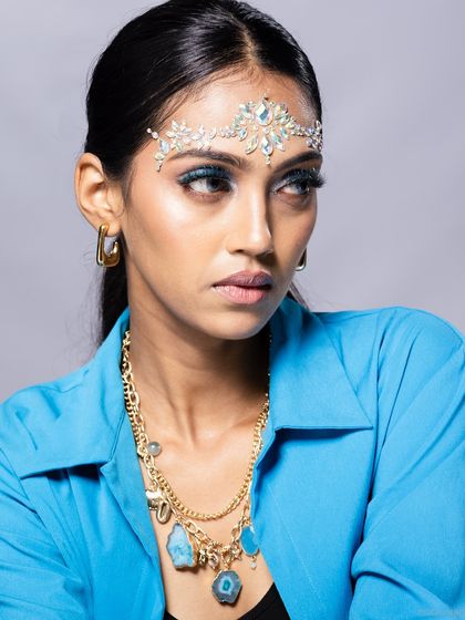 A portrait of a model wearing a crystal headpiece and layered necklace, with a thoughtful expression.