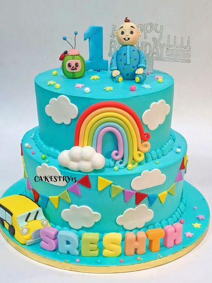 A sweet two-tier Cocomelon cake for a first birthday. The sky-blue cake is decorated with a fondant rainbow, clouds, the yellow school bus, and character toppers.