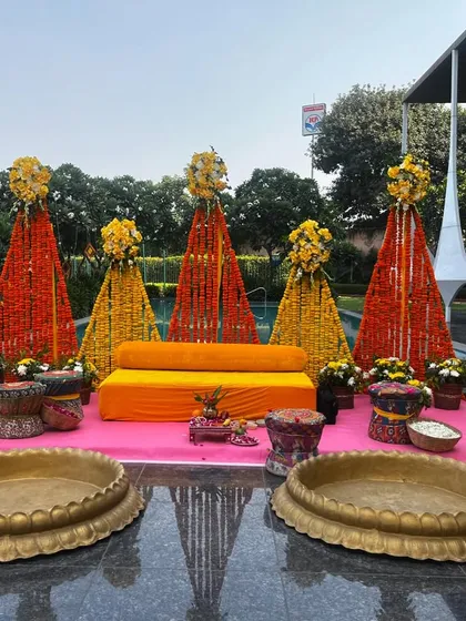 A poolside Haldi setup at Udman, Greater Noida, featuring vibrant marigold cones and a pink carpeted stage. The large brass urlis are ready for the turmeric paste, combining function with beautiful decor.