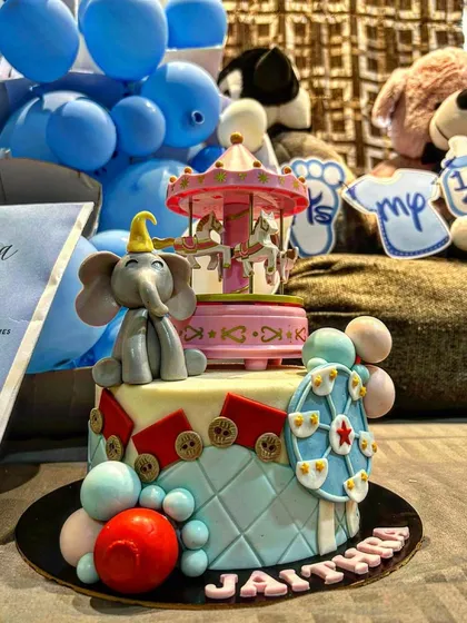 A whimsical carnival-themed cake featuring a Dumbo-style elephant, a carousel, and a Ferris wheel. The pastel colors and intricate details make it perfect for a first birthday or baby shower.