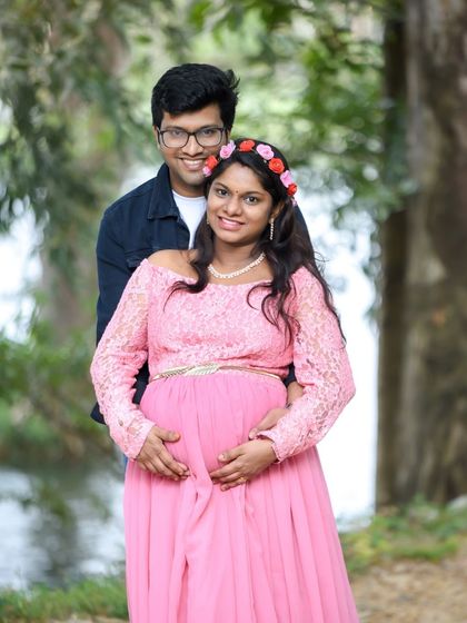 A happy and bright couple portrait by a lake. The mom-to-be is in a pink lace dress and floral headband.
