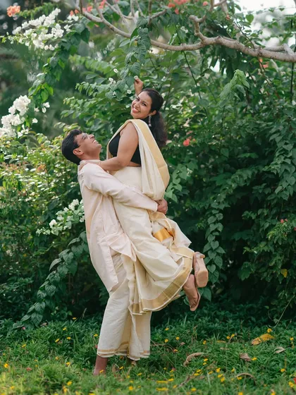 A playful and romantic moment as the groom lifts the bride, both dressed in elegant South Indian attire amidst a lush garden.