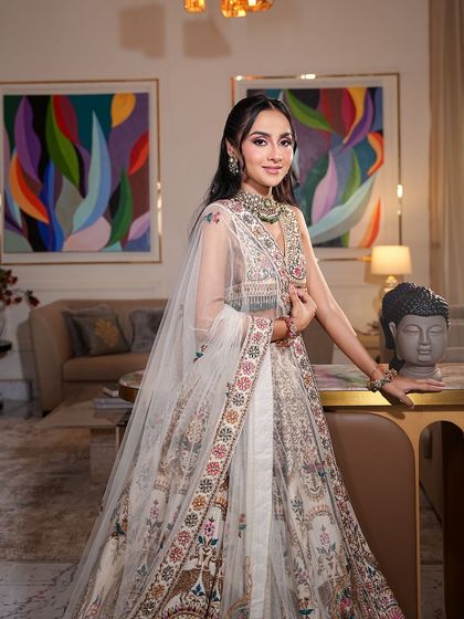 The full view of this effortlessly radiant look. The bride is seated in a modern living space, and her soft glam makeup perfectly complements the intricate, multi-colored lehenga.