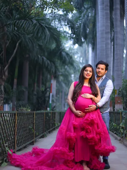 A beautiful outdoor couple's portrait on a path lined with tall trees, showcasing a full-length view of the pink maternity gown.