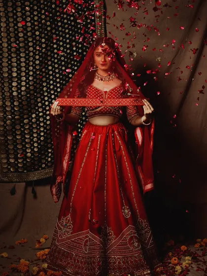 A moment of celebration in a bridal editorial. Rose petals rain down on the bride, who is dressed in a classic red lehenga. The image is designed to feel dynamic, joyful, and cinematic.