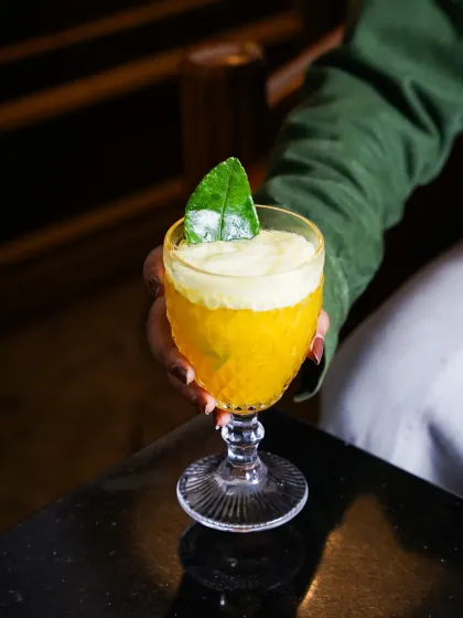 A hand in a green sleeve holds a textured glass with a frothy yellow cocktail. The focus on the hand and drink creates a personal and engaging lifestyle shot, perfect for social media.