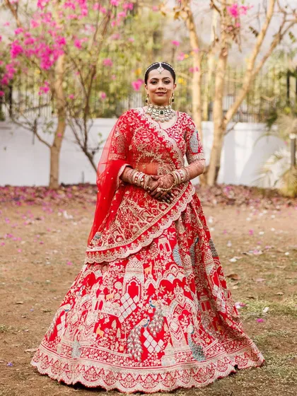 A full-length shot in a beautiful garden setting. The vibrant red of her lehenga stands out against the greenery, and her glowing makeup completes this picture-perfect bridal look.