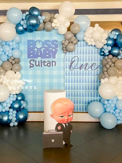 I created this stylish Boss Baby decoration for Sultan's first birthday. The design uses a blue gingham and tie-patterned backdrop, framed by a beautiful balloon arch in pastel blue, white, and metallic grey, with a character cutout as the centerpiece.