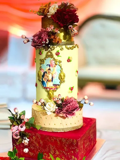 A grand, four-tiered wedding cake with an Aladdin theme. The design features a beautiful illustration of Aladdin and Jasmine, intricate gold details, and is adorned with fresh flowers.