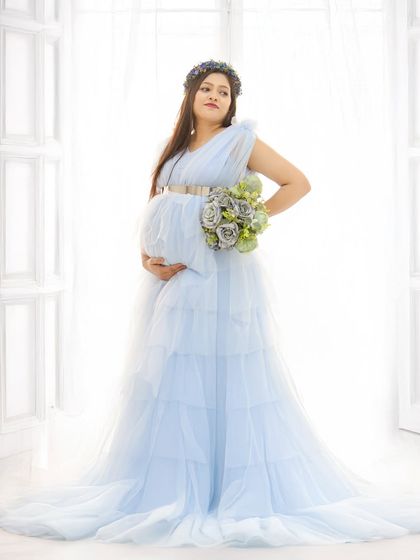 An elegant solo portrait of the mom-to-be in a light blue tulle gown. The bright, backlit window setting creates a soft, angelic glow.