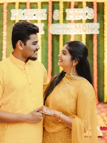 Fifi and Pratik looking at each other during their Haldi ceremony. The colorful backdrop and their matching yellow outfits create a vibrant and joyful scene.