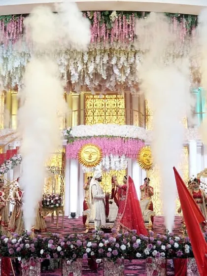 A wide shot of a grand Jaimala, showcasing the scale of the production. The couple is centered on a magnificent stage with smoke jets erupting around them, creating a breathtaking spectacle.