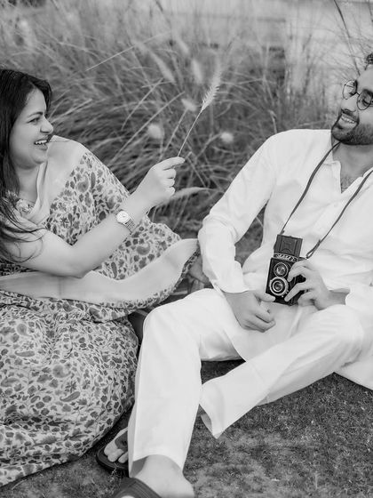 A playful and candid black and white moment. The couple shares a laugh in a field of tall grass, with the vintage camera prop adding a touch of old-school charm to their natural interaction.