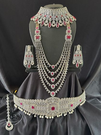 For a touch of colour, this silver-toned set with ruby-red stones is a perfect choice. The full set includes a choker, a layered necklace, and a matching vaddanam for a complete party look.