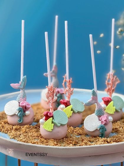 Intricately detailed cake pops with mermaid tails, seahorses, and shells, all resting on a bed of 'sand'.
