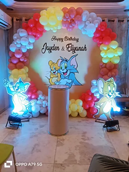 A classic Tom and Jerry theme for a joint birthday party. This setup includes a circular backdrop with the iconic duo, light-up cutouts of Tom and Jerry, and a colorful red, yellow, and white balloon arch.