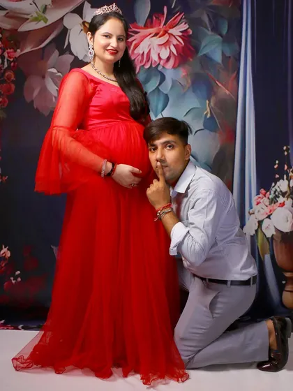 A classic couple maternity pose. The mom-to-be stands in a vibrant red gown while her partner kneels and listens to the baby bump.