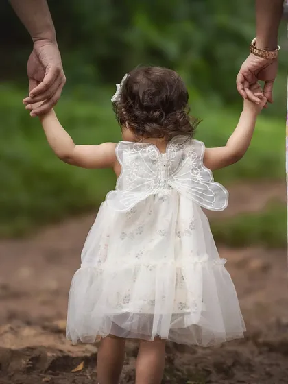 A toddler in a delicate white dress with butterfly wings holds her parents' hands as they walk through a natural path. This shot, focusing on their linked hands, symbolizes guidance, love, and the journey of family.