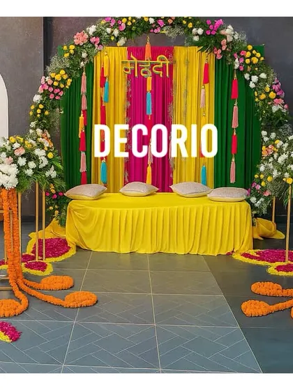 A colorful Mehndi setup featuring a backdrop of rich green and pink drapes. The comfortable floor seating with bright yellow fabric and cushions provides a perfect spot for the bride during the ceremony.