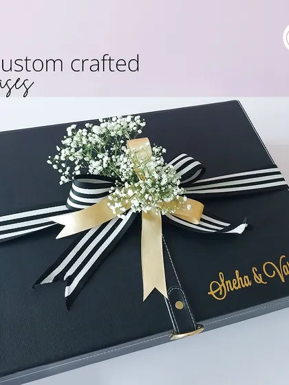 I create custom-crafted cases for a sleek, modern aesthetic. This black leatherette box features the couple's names in elegant gold script, finished with a classic striped ribbon and a touch of florals for a sophisticated look.