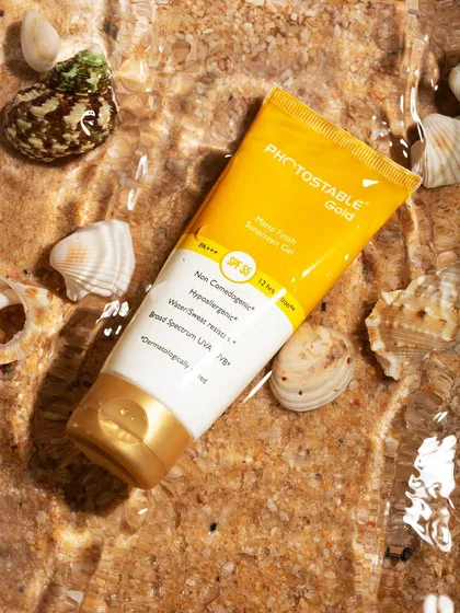 A creative shot of Photostable Gold sunscreen, styled in clear water with sand and seashells. This approach to skincare photography highlights the product's use case and creates a fresh, appealing visual for social media and ads.