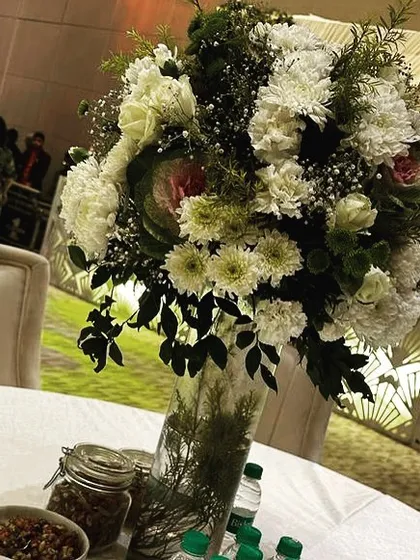 A close-up of a table centerpiece at the Roka ceremony, featuring a large bouquet of white chrysanthemums and roses in a tall glass vase.
