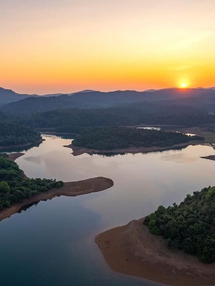 A serene sunset over a reservoir in the Western Ghats. The calm water reflects the sky and the surrounding hills, creating a peaceful and symmetrical landscape.