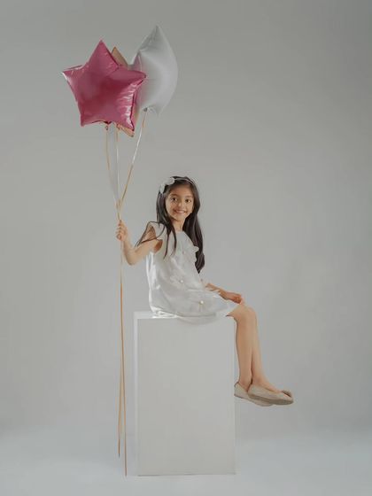 A full-length portrait of the birthday girl sitting on a white block, holding star-shaped balloons. This shows a simple yet elegant setup available in my studio.