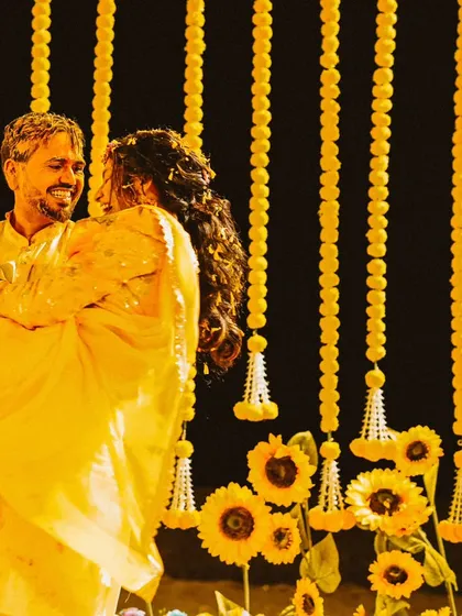 A candid moment from a Haldi ceremony, with the groom lifting the bride in his arms against a backdrop of yellow flowers.