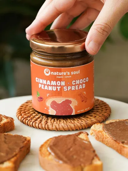 A hand opening a jar of cinnamon choco peanut spread, surrounded by toast points, creating a sense of anticipation.