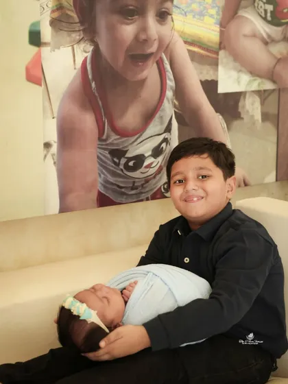 A proud big brother holds his newborn sibling for the first time. He smiles sweetly at the camera, showcasing the beginning of a special bond in this heartwarming sibling portrait.