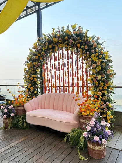 A beautiful floral arch for a Haldi or Mehendi ceremony, combining yellow flowers with hanging marigold strings and colorful tassels. The pink seashell-style sofa adds a touch of modern elegance to the vibrant, traditional setup.