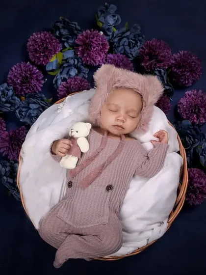 A baby in a cute bear-ear romper holds a tiny teddy bear, surrounded by a wreath of purple flowers against a deep blue background.