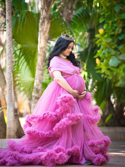 A serene outdoor portrait in a lush garden setting. The mom-to-be wears a beautiful pink ruffled gown, creating a fairytale-like image among the greenery.