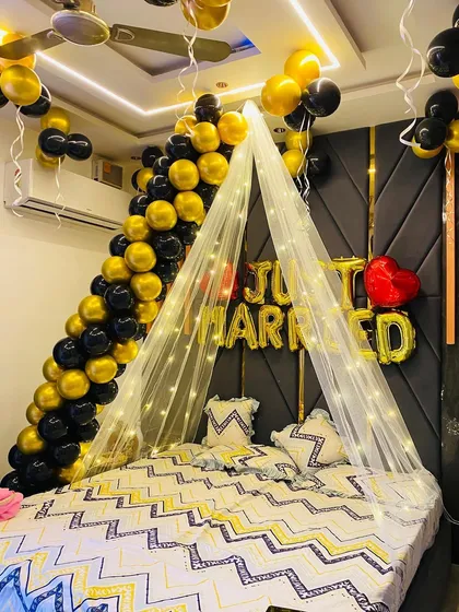 A stylish 'Just Married' room decoration using a black and gold color palette. A balloon garland flows down one side of the bed, complemented by a fairy light canopy and gold foil letters.