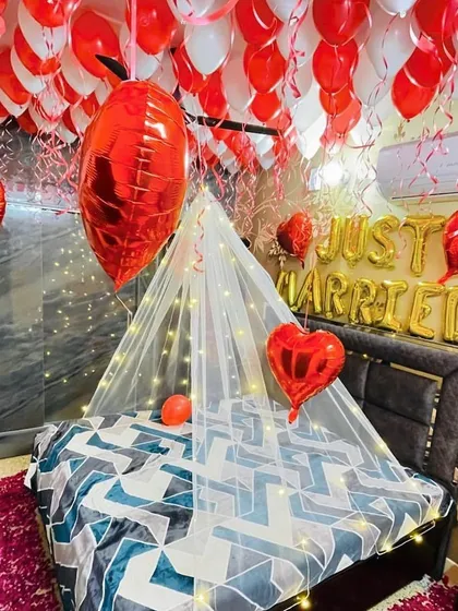 We create romantic first night or anniversary decorations for your room. This setup includes a ceiling full of red and white balloons, "Just Married" foil letters, a delicate bed canopy with fairy lights, and scattered rose petals.