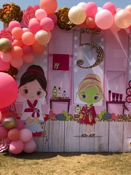 A fun and girly spa-themed 5th birthday party. The backdrop features cute cartoon characters at a spa, framed by a pink and orange balloon garland for a day of pampering and fun.