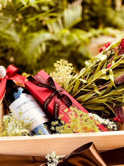 A luxurious wooden tray hamper featuring a bottle of Jacob's Creek wine and a box of Rosarte chocolates. The arrangement is complemented by a fresh bouquet of red flowers and delicate fillers, creating a classic and romantic gift.