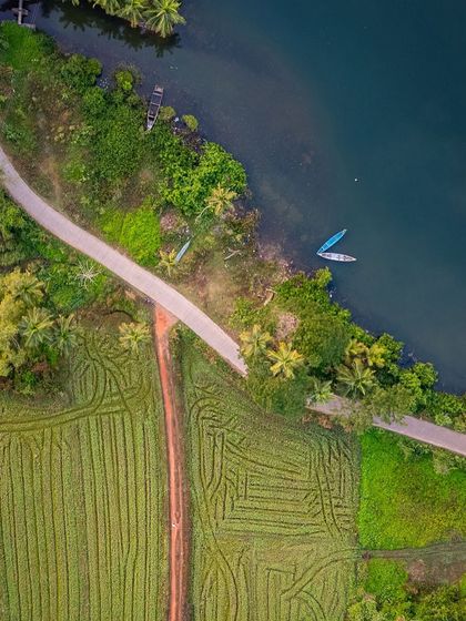 A detailed top-down drone shot showing the contrast between a dirt path, neatly plowed fields, and the deep blue water of a riverbank.