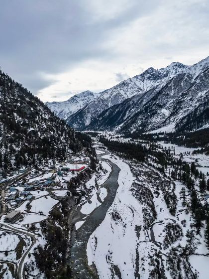 A sweeping aerial view of a snow-covered valley in Spiti, showing the river carving its path through the winter landscape. This is a classic example of Himalayan drone photography, capturing the vastness of the region.
